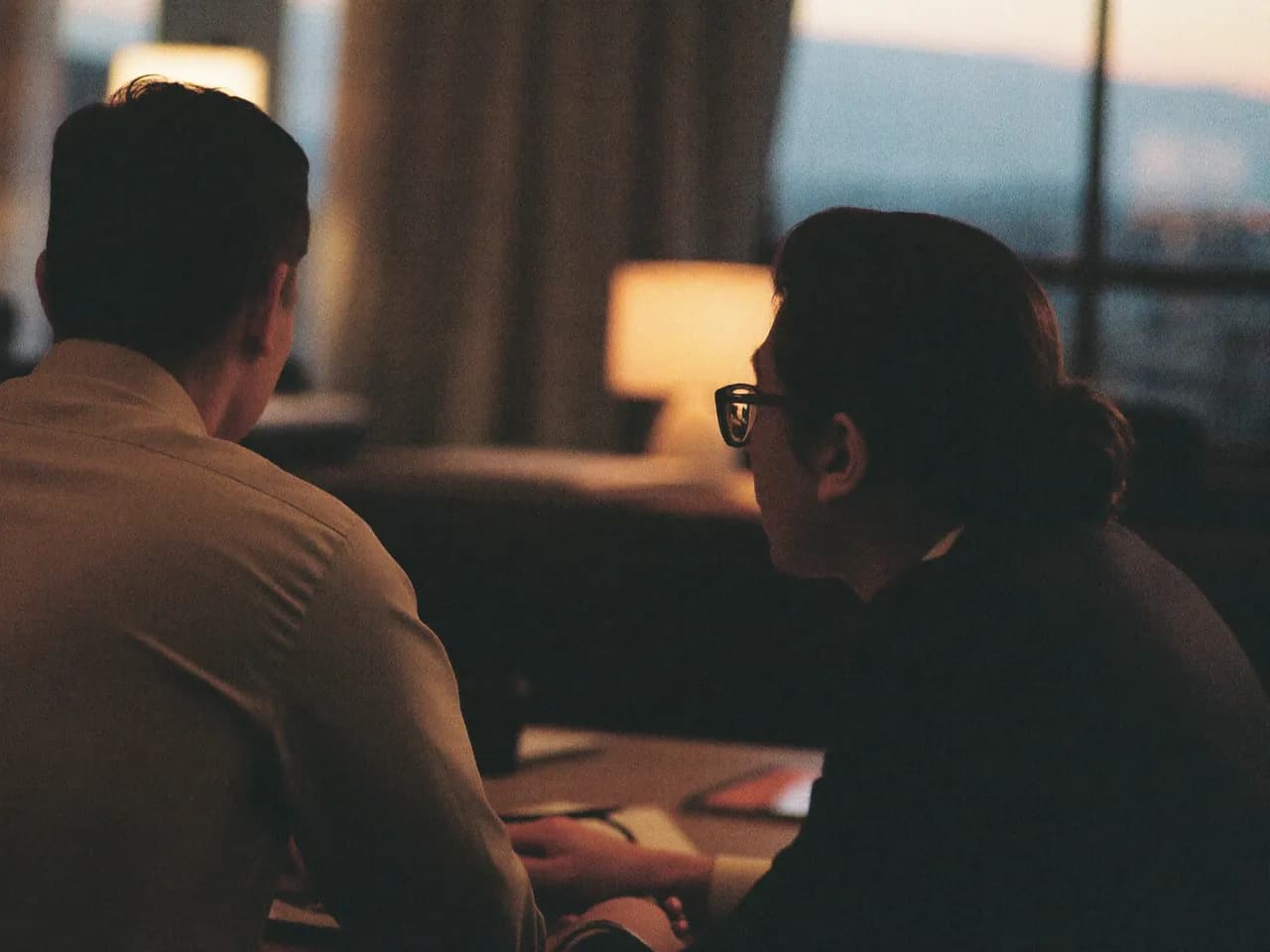 Two colleagues at a desk at dusk, seen from behind, city light beyond the window